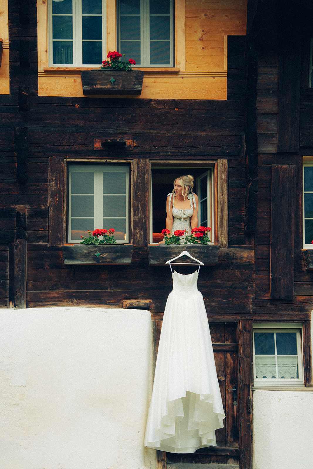 A cinematic photo of a bride beside her wedding dress that is hanging from a cabin's window in Switzerland.